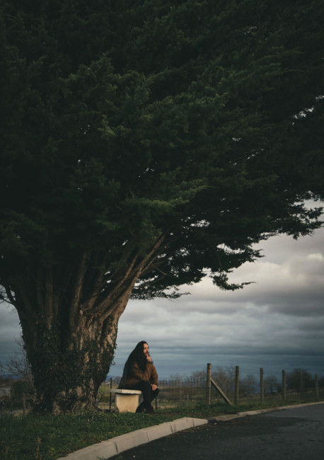 Au bord d'une petite route, une personne songeuse est assise sur un banc en pierre. Derrière elle, un arbre au tronc très épais. Le feuillage est immense et recouvre la moitié du ciel.
