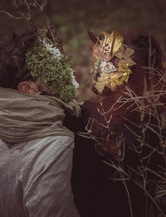 Vues de profil, deux personnes se font face. Leurs vêtements se frottent. Leurs visages sont recouverts par des masques mystérieux, faits de lichen vert et de feuilles d'automne.